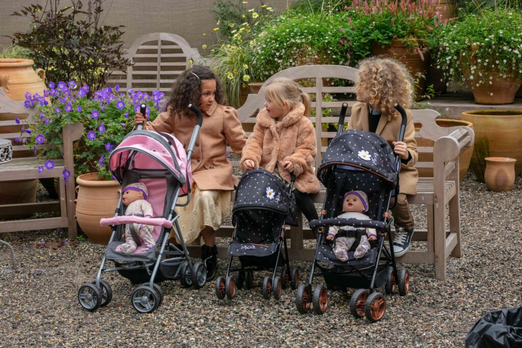 A group of children sat on an outdoors bench with their dolls pushchairs. The image is illustrating a dolls pram buyers guide.