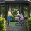 Daisy Chain Connect Dolls Pram in Classic Pink fabric shown on a side angle with the handle to the left side. Hood is up and is pink with accents of Silver metal with a black and pink flower rosette and an with an embroidered Daisy Chain logo in silver. Wheels are black and Silver with a shopping basket at bottom of pushchair which is black. The frame of the pushchair is silver. Girl with long brown hair standing behind the pushchair in a blue denim skirt and yellow cardigan.She is stood infront of a pagoda on a cobblestone bridge.