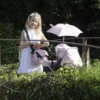 Daisy Chain Dolls Pram Accessory Pack in Classic Pink fabric. Photo shows a girl with long blonde hair stood behind the connect pram with a shoulder bag over her and a pink parasol on the pram. She is stood on a bridge with hedges round her.