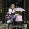 Daisy Chain Dolls Pram Accessory Pack in Classic Pink fabric. Photo shows a girl with long blonde hair stood behind the connect pram with a shoulder bag over her and a pink parasol on the pram. She is stood on a bridge in front of a pagoda.