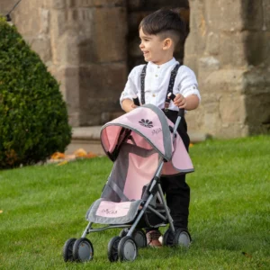 Daisy Chain Little Zipp Dolls Pushchair in Classic Pink fabric shown on an angle with the handles on the right side. Hood is down and is pink at the front with accents of grey fabric with a black and pink flower rosette and an with an embroidered Daisy Chain logo in silver. Wheels are black and Silver with a shopping basket at bottom of pushchair which is black. The frame of the pushchair is silver. Little boy with dark brown hair standing behind the pushchair in a white shirt and trousers with braces. Stood on grass in front of a church building.