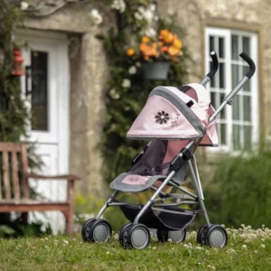 Daisy Chain Zipp Max Dolls Pushchair in Classic Pink fabric shown on an angle with the handles on the right side. Hood is partly up and is mainly pink with accents of grey fabric with a black and pink flower rosette. Seat is in pink and grey fabric. Wheels are black and shopping basket at bottom of pushchair is black. The pram is on the grass with a house in the background. The house has a white door with a bench on the left side and an orange hanging basket on the other side of the door.