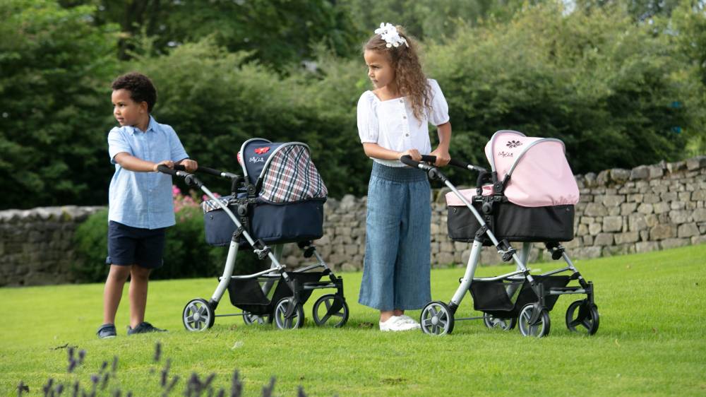an image of two children playing with dolls prams and doll car seats. The children are outdoors stood on a grassy area.