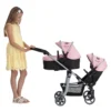 Daisy Chain Pinnacle Double Dolls Pram in Pink fabric for ages 6-13 years old. The pram has a silver frame with black accents. Cot has a black fabric body and pushchair seat has a black body. Both the cot and seat have hoods which are pink with a black and pink daisy that is removable. The cot has a pink apron. Girl stood next to the pram with a yellow dress on.