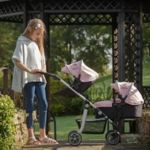 Daisy Chain Pinnacle Double Dolls Pram in Pink fabric for ages 6-13 years old. The pram has a silver frame with black accents. Cot has a black fabric body and pushchair seat has a black body. Both the cot and seat have hoods which are pink with a black and pink daisy that is removable. The cot has a pink apron. Girl stood next to the pram with summer clothes on.