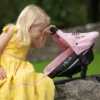 Daisy Chain Unity High Chair/Car Seat in Classic Pink fabric. Girl with shoulder length blonde hair in a yellow summer dress sitting on a dry-stone wall. She is holding the handle of the car seat and looking at her doll.