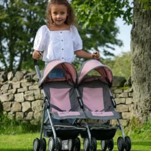 Daisy Chain Zipp Twin Max Dolls Pushchair in Classic pink fabric shown on an angle with the handles on the left side. Hoods are partly up and are mainly pink with accents of grey fabric with a black and pink flower rosette. The pushchair has 2 seats side-by-side in the pink fabric with accents of grey and an embroidered Daisy Chain logo in silver. The wheels are black and the shopping baskets at bottom of pushchair are black. A young girl with curly brown hair is standing behind the pushchair and is wearing a white top and blue denim jeans, she is stood in front of a dry stone wall and a big tree.