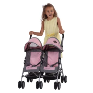 Daisy Chain Zipp Twin Max Dolls Pushchair in Classic pink fabric shown on an angle with the handles on the left side. Hoods are partly up and are mainly pink with accents of grey fabric with a black and pink flower rosette. The pushchair has 2 seats side-by-side in the pink fabric with accents of grey and an embroidered Daisy Chain logo in silver. The wheels are black and the shopping baskets at bottom of pushchair are black. A young girl with curly brown hair is standing behind the pushchair and is wearing a yellow summer dress.