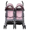 Daisy Chain Zipp Twin Max Dolls Pushchair in Classic Pink fabric. Shown front on. The pushchair has 2 seats side-by-side in the pink fabric with accents of grey and an embroidered Daisy Chain logo in silver. The pushchair has two hoods partly up and are mainly pink with accents of grey with a black and pink flower rosette. The wheels are black. The frame is silver and the two shopping baskets at bottom of the pushchair are black.