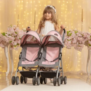 Daisy Chain Zipp Twin Max Dolls Pushchair in Classic pink fabric shown on an angle with the handles on the left side. Hoods are partly up and are mainly pink with accents of grey fabric with a black and pink flower rosette. The pushchair has 2 seats side-by-side in the pink fabric with accents of grey and an embroidered Daisy Chain logo in silver. The wheels are black and the shopping baskets at bottom of pushchair are black. A Young girl with blonde hair is stood behind the pushchair in a cream dress with fairy lights and pink flowers in the background.