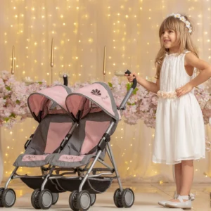 Daisy Chain Zipp Twin Max Dolls Pushchair in Classic pink fabric shown on an angle with the handles on the left side. Hoods are partly up and are mainly pink with accents of grey fabric with a black and pink flower rosette. The pushchair has 2 seats side-by-side in the pink fabric with accents of grey and an embroidered Daisy Chain logo in silver. The wheels are black and the shopping baskets at bottom of pushchair are black. A Young girl with blonde hair is stood next to the pushchair in a cream dress with fairy lights and pink flowers in the background.