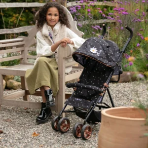 Daisy Chain Zipp Zenith Dolls Pushchair in Limited Edition Twilight fabric shown head on with the handles at the back. Hood is black with little daisies on the fabric with a white daisy rossette with a yellow centre on the side of the hood. Seat is black with daisies on it. It has a rose gold trim with an embroidered white Daisy Chain logo. The pushchair features a black bumper bar that can be raised up and down to make it easy to get your doll in and out of the pushchair. Wheels are rose gold with black tyres. With shopping basket at bottom of pushchair which is black. The frame of the pushchair is black with accents of rose gold. Girl with long dark hair wearing a fluffy cream gillet, green skirt and black boots she is sat next to the pushchair on a bench. Set on a gravel path in front of flowers and green shrubs.