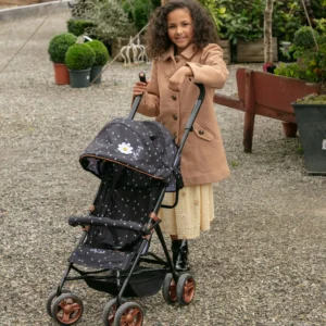 Daisy Chain Zipp Zenith Dolls Pushchair in Limited Edition Twilight fabric shown head on with the handles at the back. Hood is black with little daisies on the fabric with a white daisy rossette with a yellow centre on the side of the hood. Seat is black with daisies on it. It has a rose gold trim with an embroidered white Daisy Chain logo. The pushchair features a black bumper bar that can be raised up and down to make it easy to get your doll in and out of the pushchair. Wheels are rose gold with black tyres. With shopping basket at bottom of pushchair which is black. The frame of the pushchair is black with accents of rose gold. Girl with long dark hair wearing a camcel coloured coat, yellow dress and black boots she is stoof up behind the pushchair. Set on a gravel path in front of flowers and green shrubs.