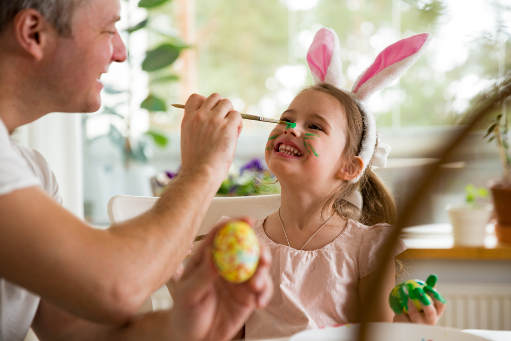 A father and daughter celebrating Easter, painting eggs with brush. Happy family smiling and laughing, drawing on face. Cute little girl in bunny ears preparing the holiday.