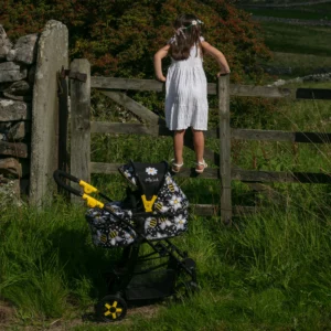 Girl climbing on a field gate behind the Daisy Chain Connect Dolls Pram in bumblebee fabric, black frame and yellow accents. Pram positioned on the grass.