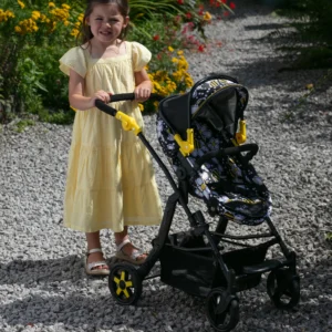 Girl in yellow dress pushing the Daisy Chain Connect dolls pram - in pushchair mode - with bumblebee fabric, black frame and yellow accents. Gravel pathway with flowers in the background.