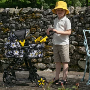 Boy in yellow hat pushing the Daisy Chain Connect dolls pram with bumblebee fabric, black frame and yellow accents. Dry stone wall in the background and chair on right mostly out of shot.