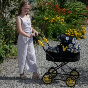 Daisy Chain Destiny Travel System Dolls Pram in Bumblebee fabric. The pram has a black frame, cot has a black quilted fabric body with an apron on top in bumblebee fabric and hood is half black and half bumblebee fabric. The pram has yellow wheels with black tyres. Girl standing behind the pushchair in a striped jumpsuit stood on gravel with flowers and shrubs in the background.