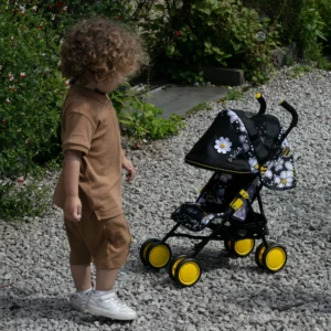 Daisy Chain Little Zipp Dolls Pushchair on gravel with a black and bumblebee fabric print and yellow wheels. Boy on left with light curly hair and light brown top and bottoms with white trainers. Shrubs and plants in background.