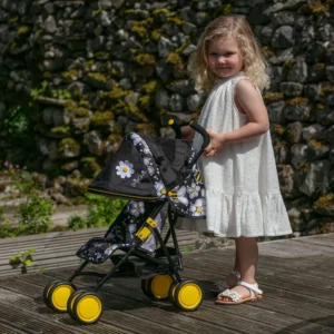 Daisy Chain Little Zipp Dolls Pushchair has black and bumblebee fabric print and yellow wheels. Girl standing behind the pushchair in white knee length dress and white sandals. She has blond curly hair. They are stood on wood decking with a dry stone wall in the background.