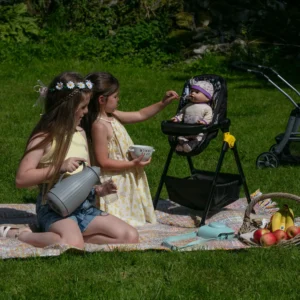 Girl with long brown hair is feeding her doll which is sat in the Daisy Chain High Chain in bumblebee fabric.. In a picnic setting with another girl to her left pouring a drink.