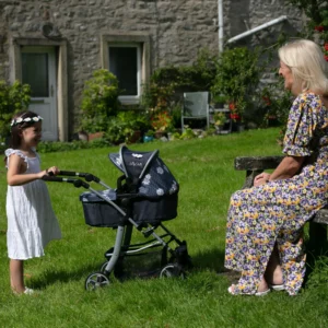 Daisy Chain Connect Dolls Pram in Daisy Dot fabric shown side on with handle on the left. Hood is up and is mainly navy blue with accents of daisy's and white polka dots Silver metal with a white and yellow flower rosette and an with an embroidered Daisy Chain logo in white. Wheels are black and Silver with a shopping basket at bottom of pushchair which is black. The frame of the pushchair is silver. A young girl with long dark hair is standing behind the pram looking at her granny, they are smiling at each other.