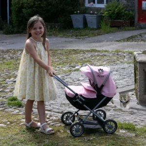 Daisy Chain Pocket Pram in Classic Pink - For ages 18 months - 3 years. Silver frame, black cot and pink hood. Girl standing next to the pram in a summer dress.