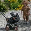 Daisy Chain Pocket Pram in Limited Edition Twilight - For ages 18 months - 3 years. Black frame with rose gold accents, black cot and limited edition twilight hood. Boy walking towards it in brown top and bottoms.